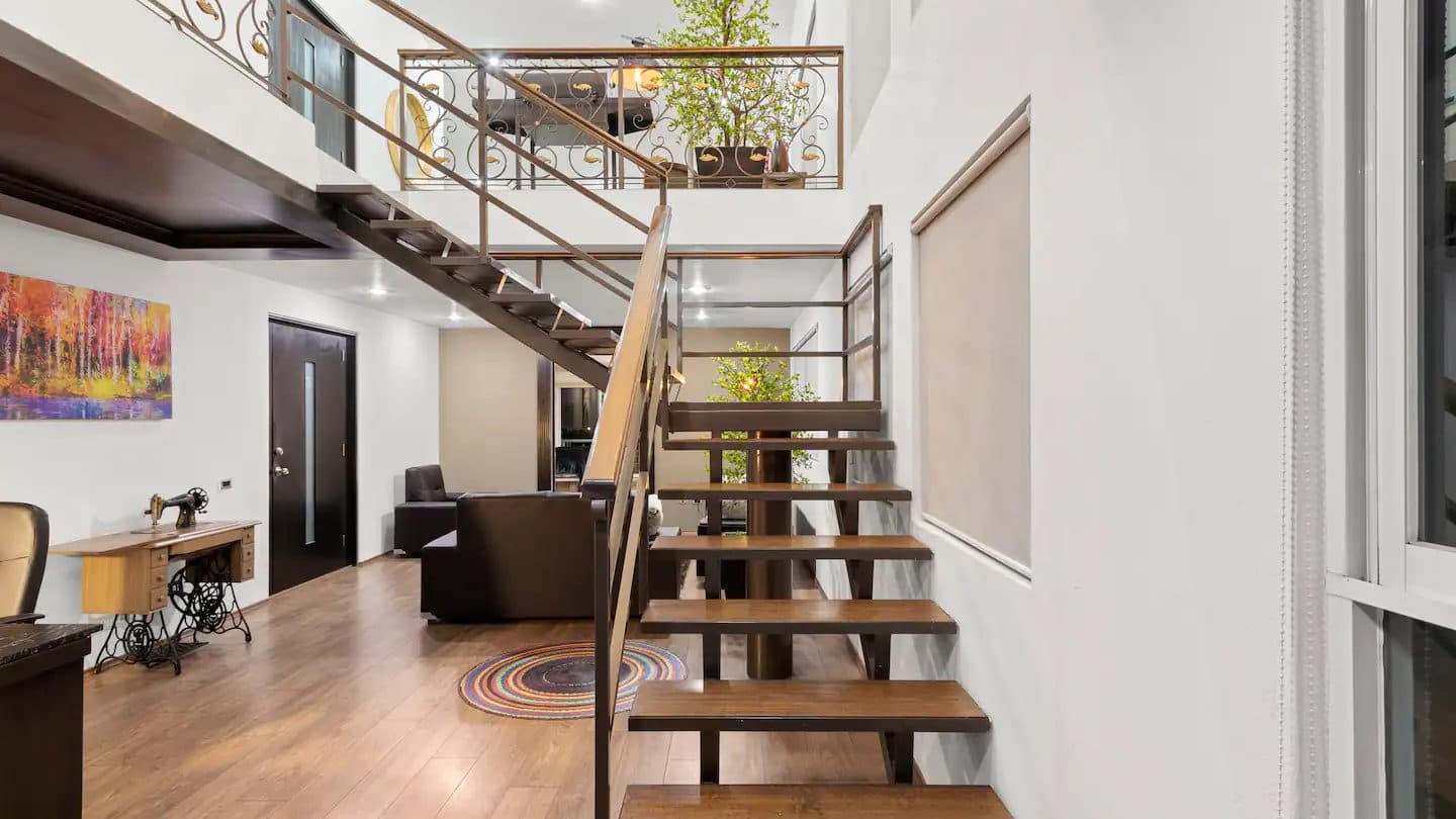 Escalera de peldaños de madera con barandal metálico en un interior de doble altura.