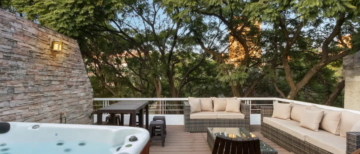 Terraza de madera con jacuzzi, salas exteriores y árboles al atardecer.