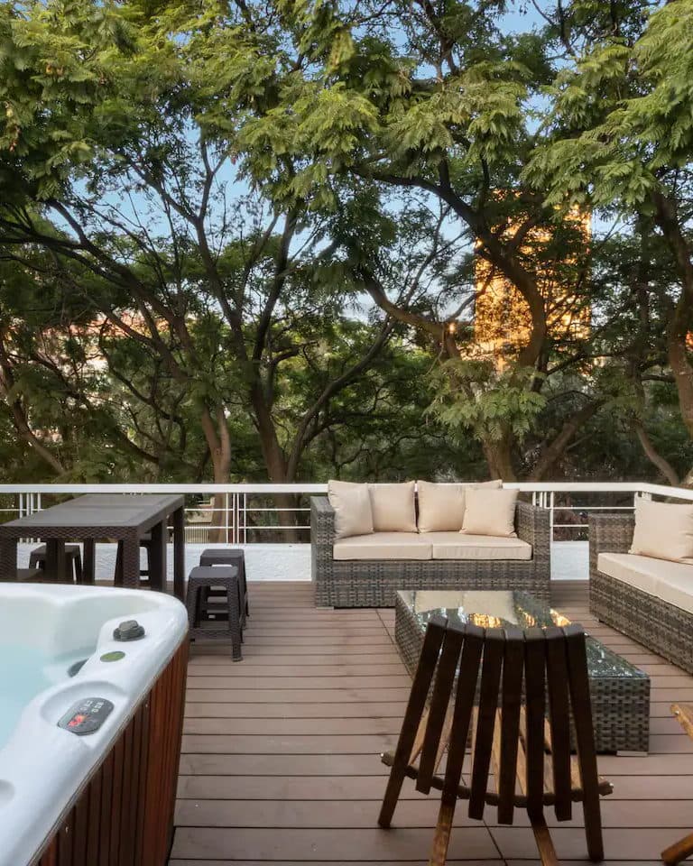 Terraza de madera con jacuzzi, salas exteriores y árboles al atardecer.